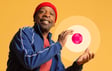 Smiling man in a bright blue jacket and red beanie gestures with one hand towards a pink and white circular graphic set against a warm yellow background.